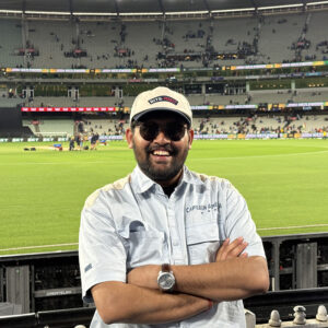 Smiling student in a white shirt and cap standing in a stadium with a crowd in the background.